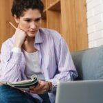 A woman sitting with her legs on the sofa in front of a laptop and writing something with her hand on her chin in the room
