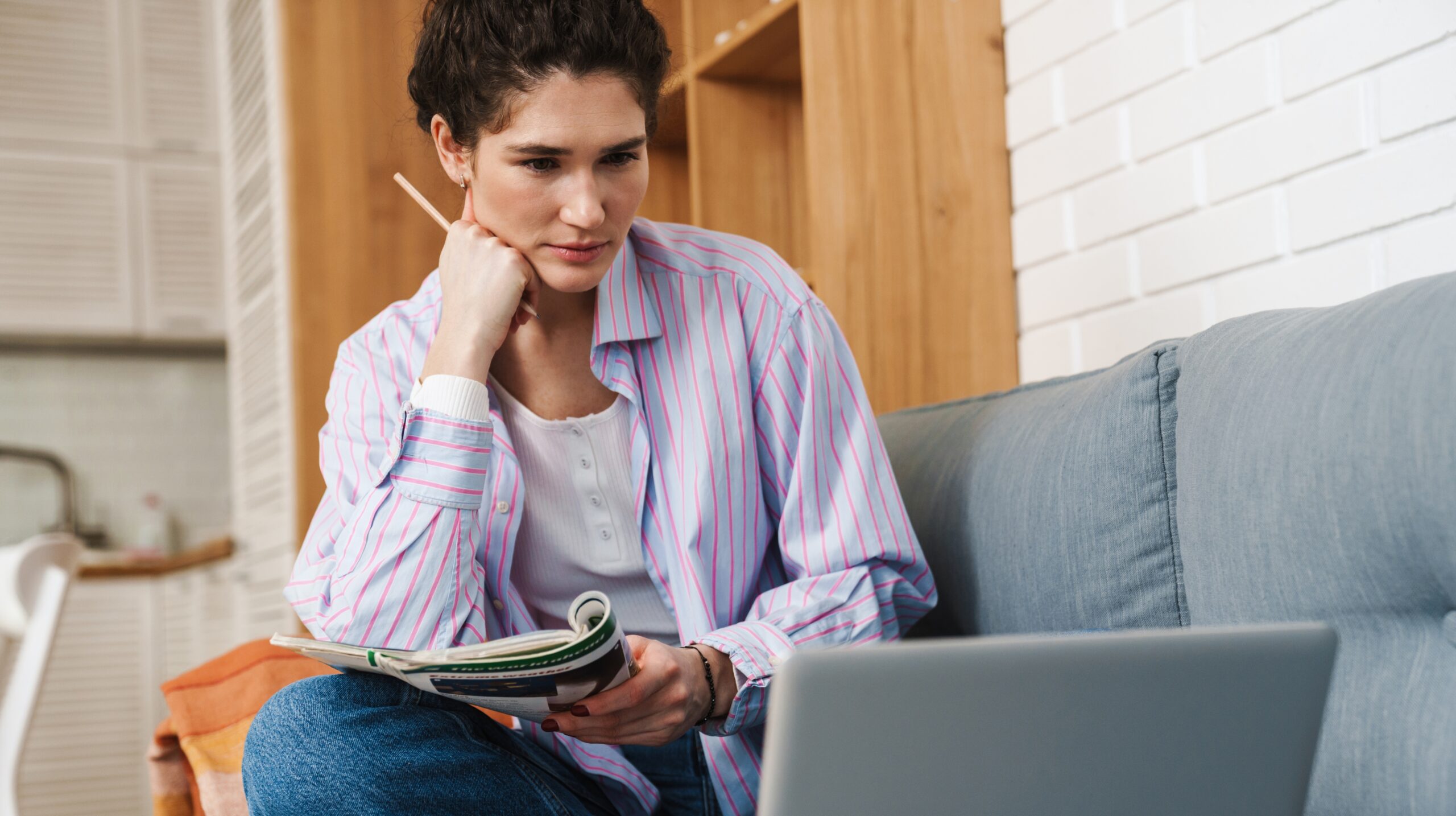 A woman sitting with her legs on the sofa in front of a laptop and writing something with her hand on her chin in the room