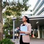 A mid-30s internationally trained nurse with dark hair in a ponytail stands at the entrance of a modern Australian hospital in soft morning light. She wears a beige blazer, holds a blue folder and passport, and looks hopefully toward the building. Eucalyptus trees and Australian flags are visible in the background. The image has a warm, documentary-style feel.