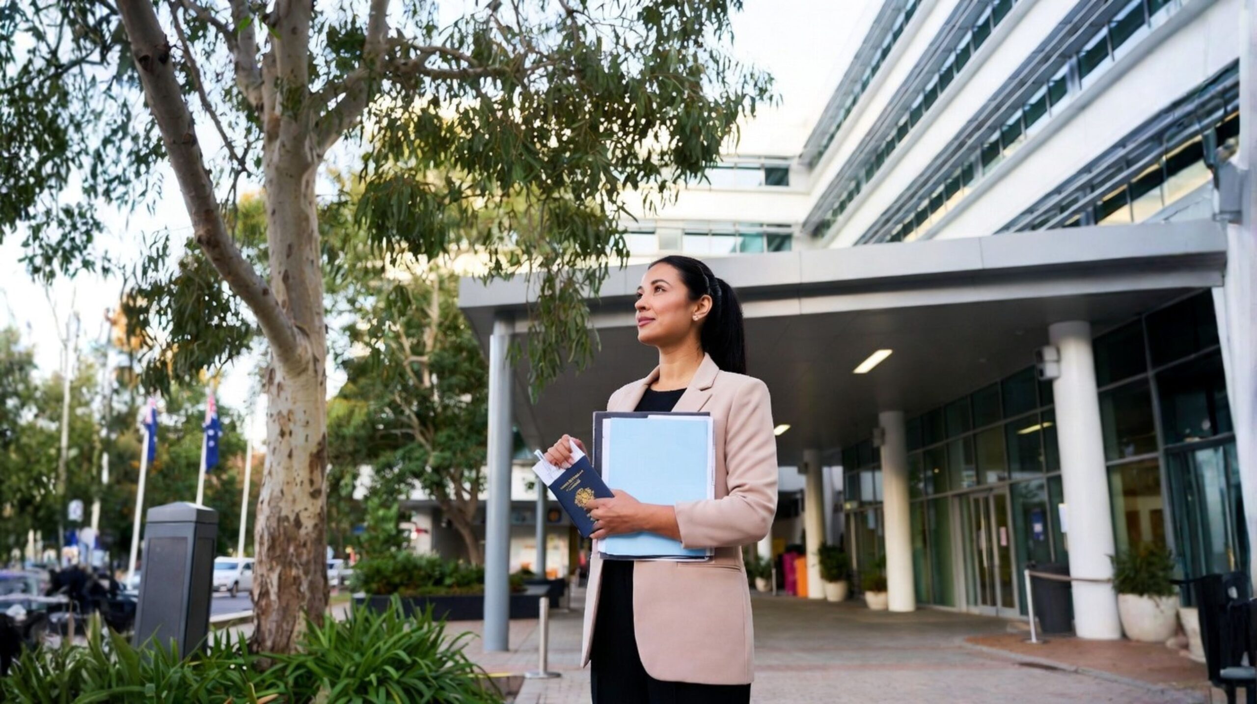 A mid-30s internationally trained nurse with dark hair in a ponytail stands at the entrance of a modern Australian hospital in soft morning light. She wears a beige blazer, holds a blue folder and passport, and looks hopefully toward the building. Eucalyptus trees and Australian flags are visible in the background. The image has a warm, documentary-style feel.