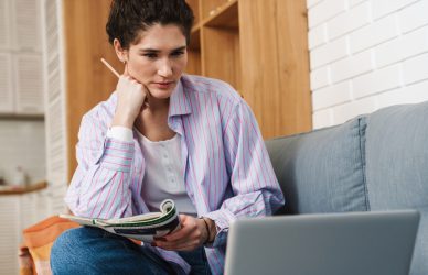 A woman sitting with her legs on the sofa in front of a laptop and writing something with her hand on her chin in the room