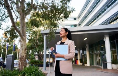 A mid-30s internationally trained nurse with dark hair in a ponytail stands at the entrance of a modern Australian hospital in soft morning light. She wears a beige blazer, holds a blue folder and passport, and looks hopefully toward the building. Eucalyptus trees and Australian flags are visible in the background. The image has a warm, documentary-style feel.